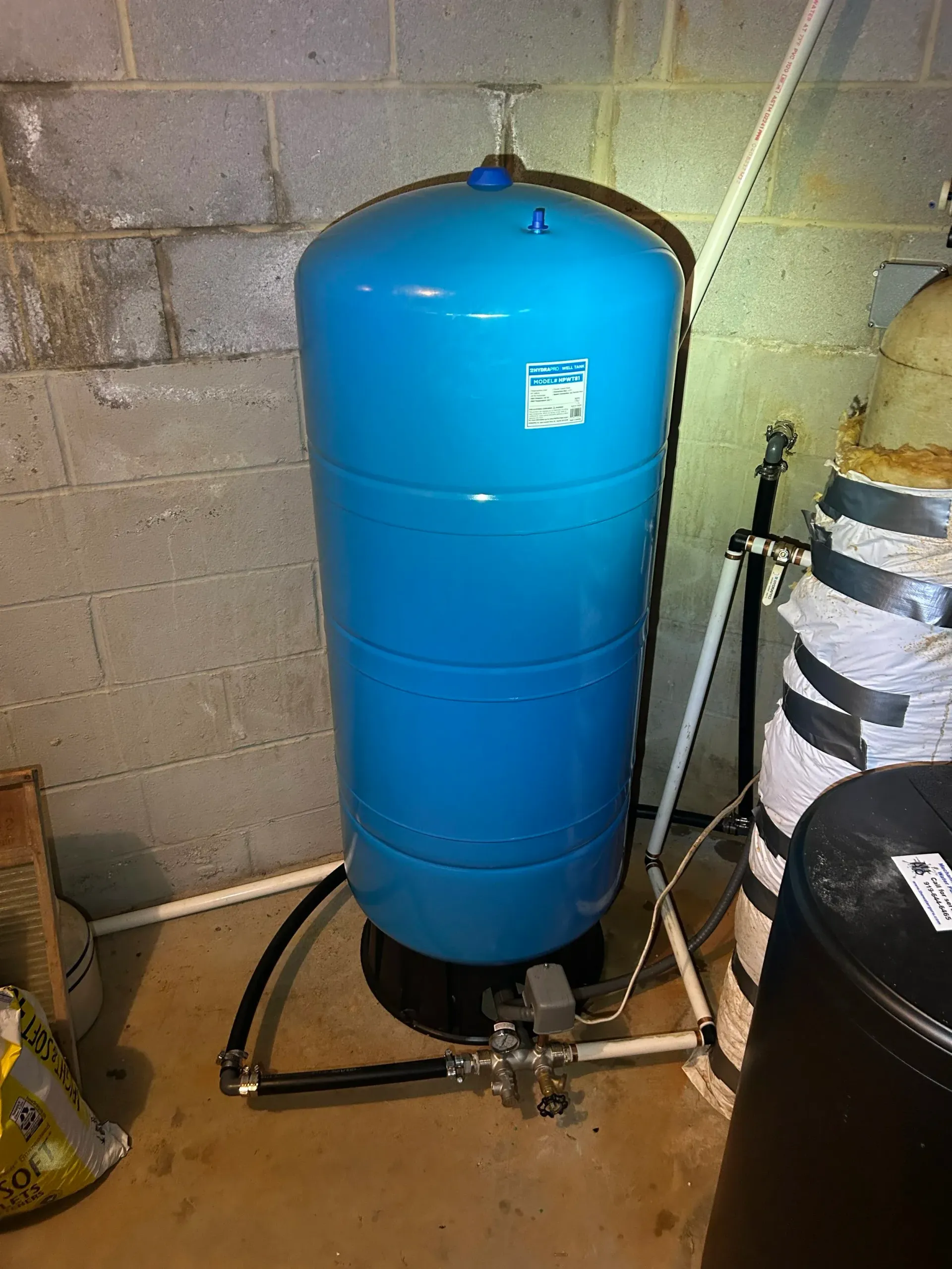 Blue water pressure tank in a basement, connected to pipes near a cinder block wall.