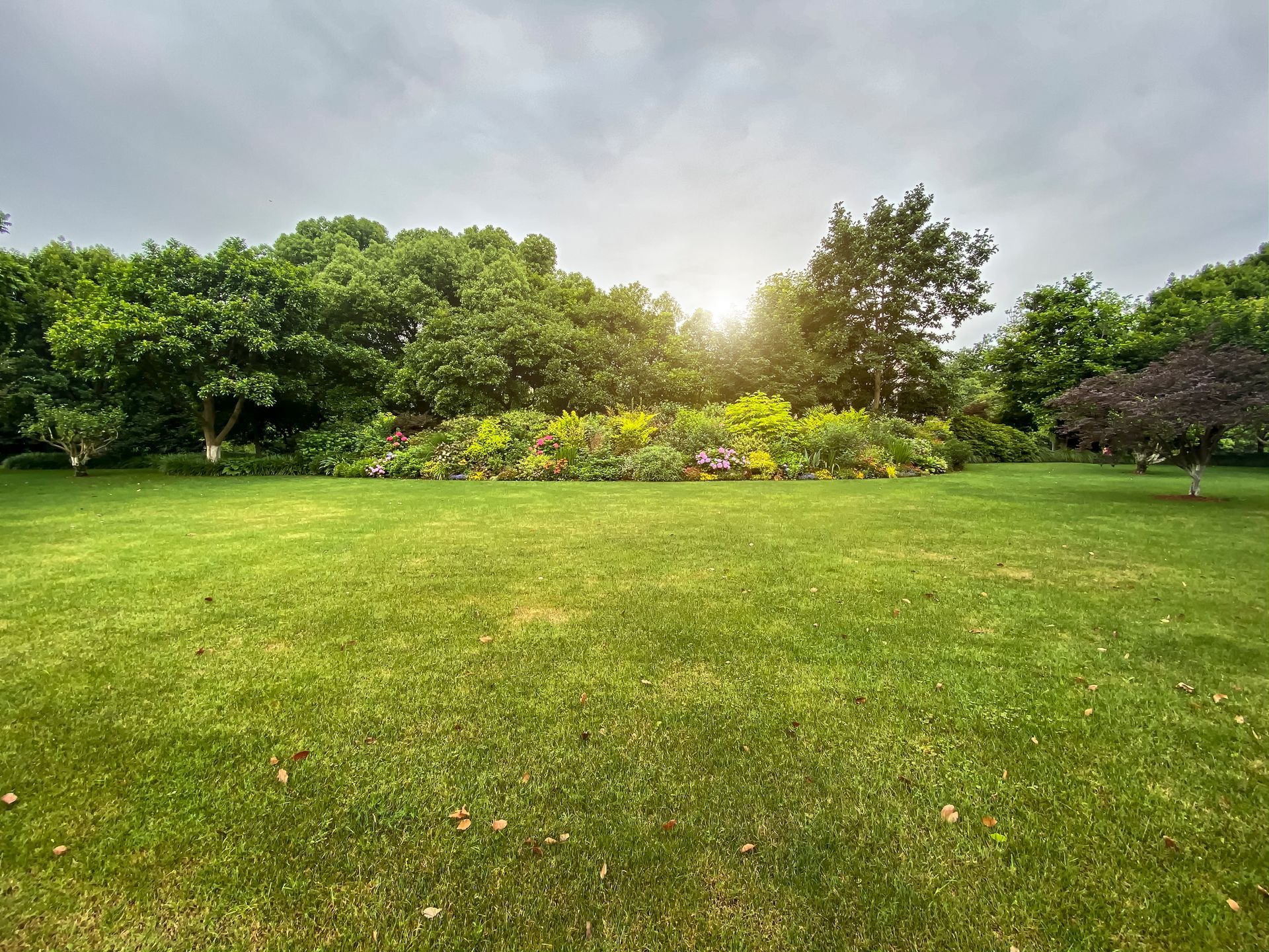 Lush green lawn with a vibrant flower bed and trees under a cloudy sky.