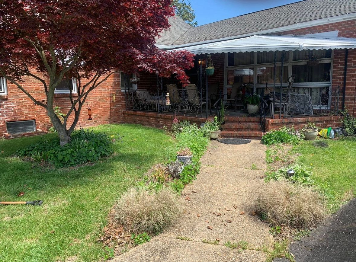 Red brick house with a gravel path leading to a covered porch with chairs. Green grass and red tree.
