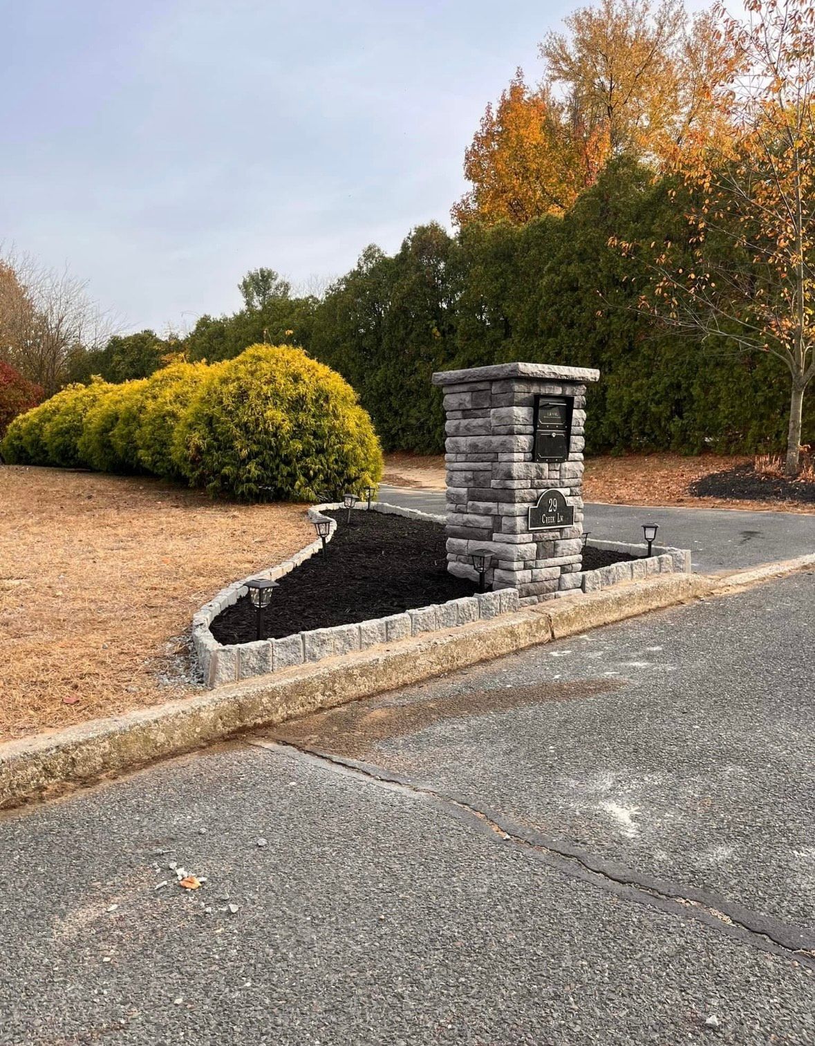Stone entrance pillar with black mulch bed, asphalt road, and green/yellow bushes.