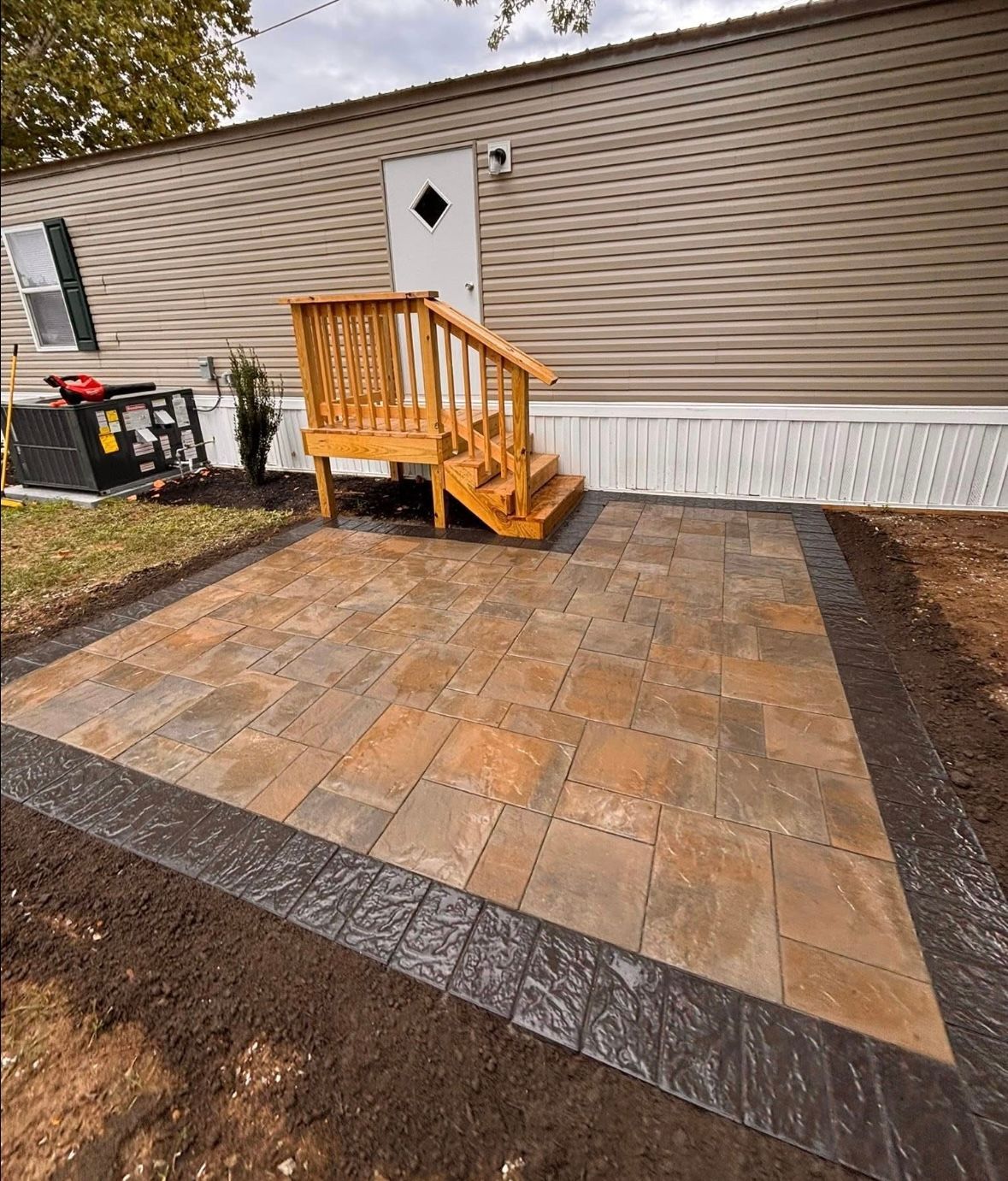 Patio with brown and gray pavers in front of a tan mobile home with wooden steps.