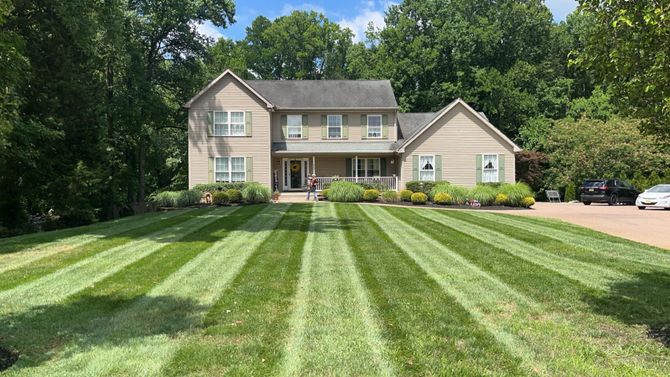 Two people mowing grass; one uses a mower, the other a leaf blower, on a grassy lawn next to a sidewalk. Two people mowing grass; one uses a mower, the other a leaf blower, on a grassy lawn next to a sidewalk.