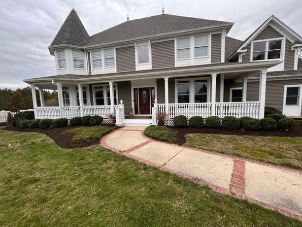 Large, brown-sided house with a porch. A brick pathway leads to the front door. Green shrubs line the porch. Large, brown-sided house with a porch. A brick pathway leads to the front door. Green shrubs line the porch.