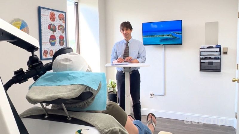 A healthcare professional stands at a desk in a medical office, facing a patient on a treatment table.
