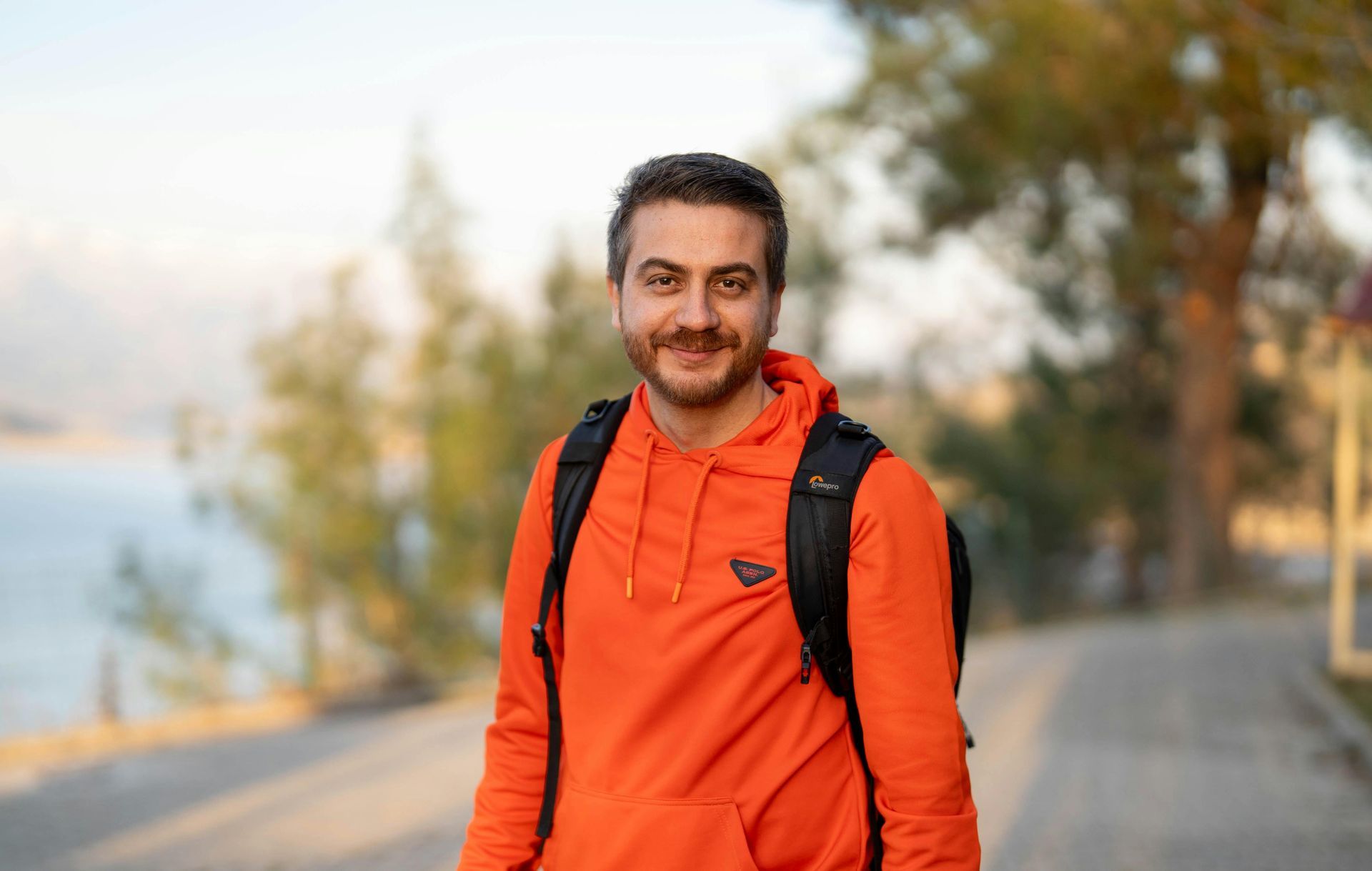 A man wearing an orange hoodie and a backpack is standing in front of a body of water.