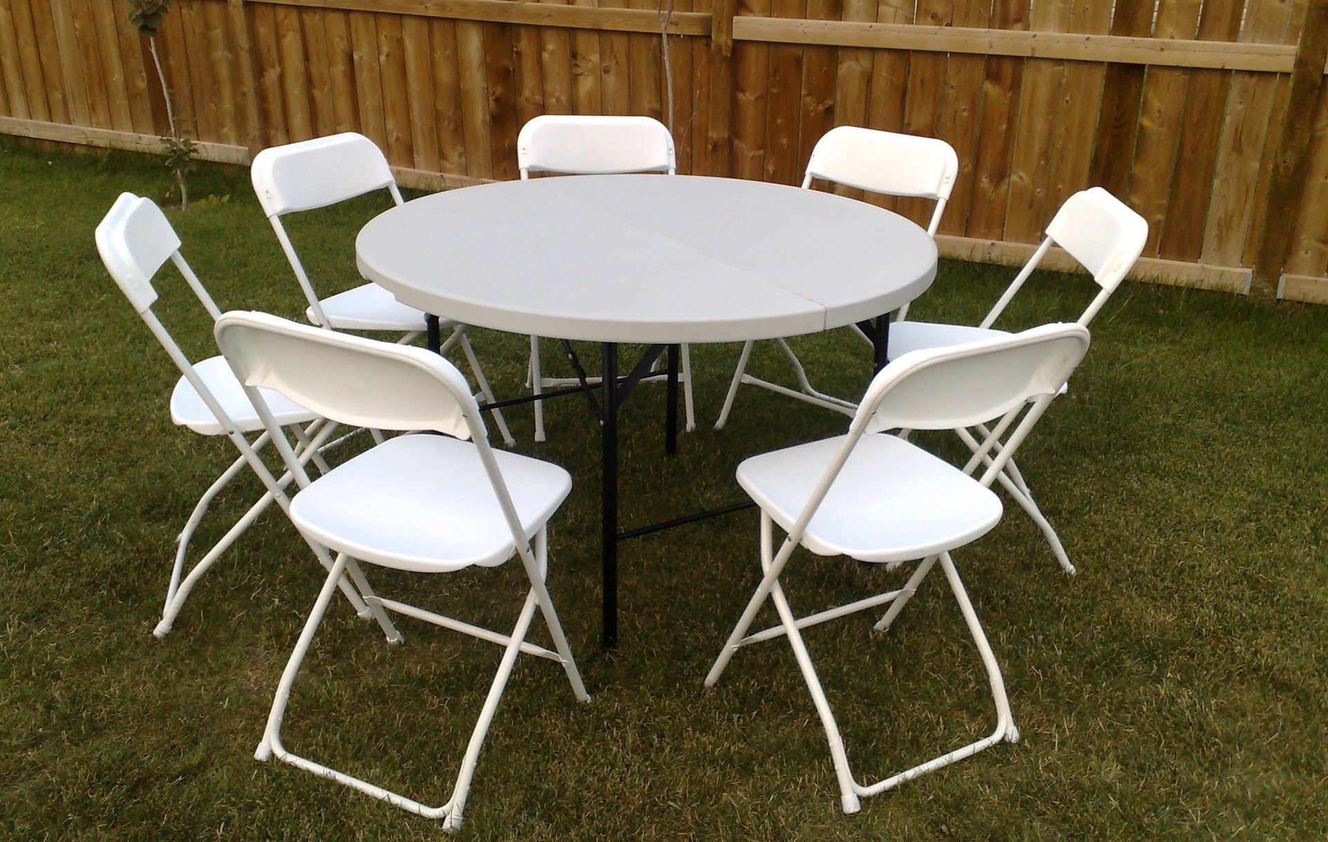 Round white table with six white folding chairs on grass in front of a wooden fence