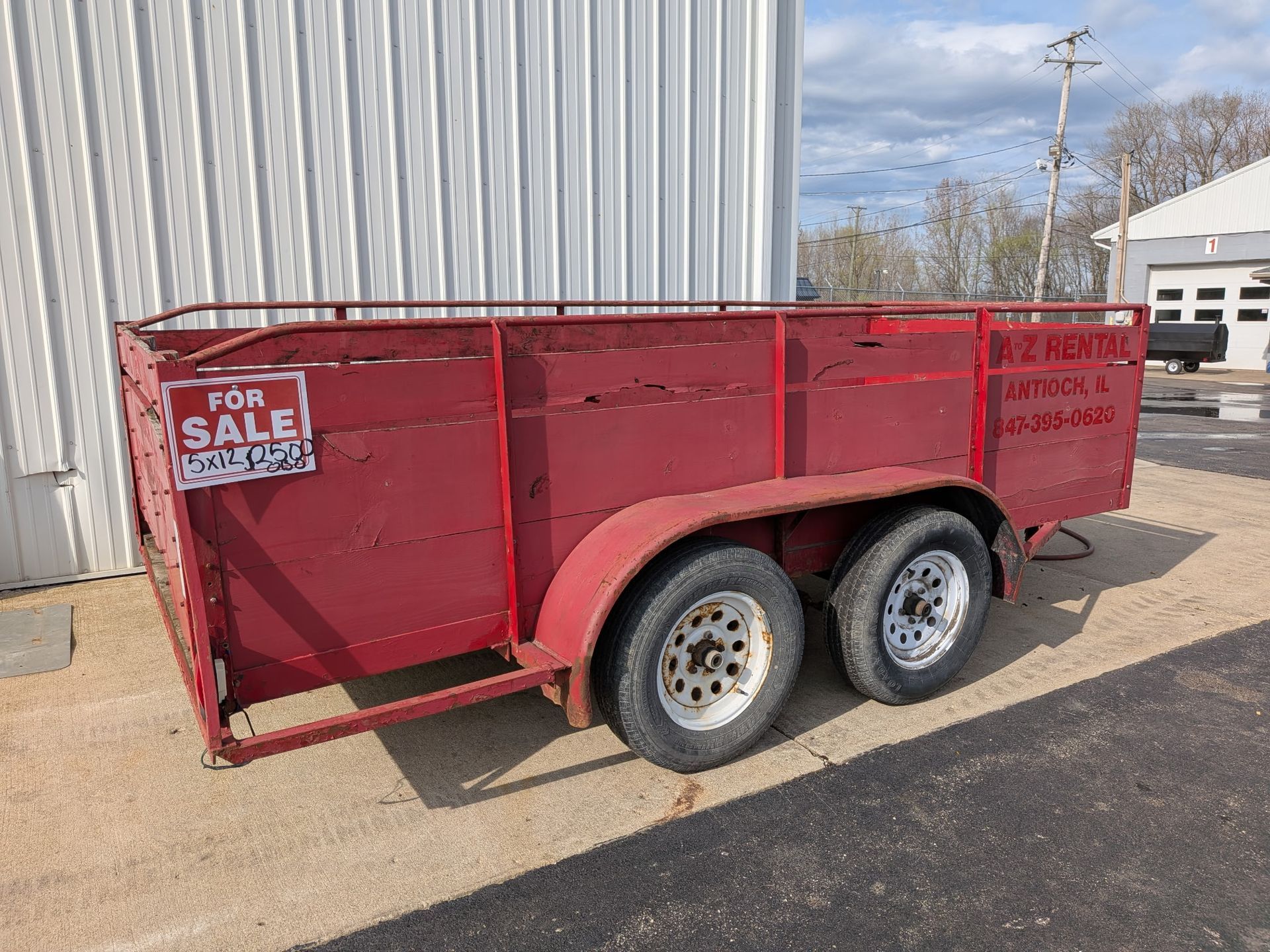 Red dual-axle utility trailer with a “For Sale” sign parked beside a metal building.