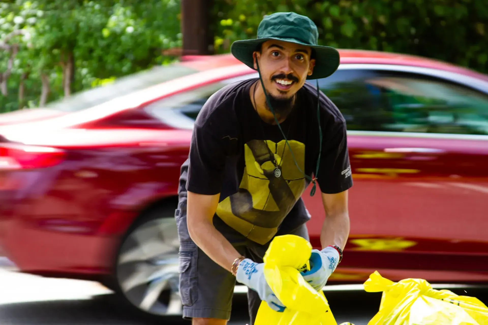 A man wearing a hat is holding a yellow bag in front of a red car.