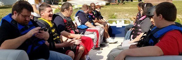 People wearing life vests on a boat. Some laughing, one looks at his phone. Outdoors, sunny.