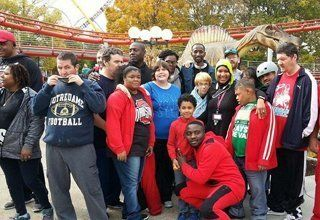 Group of people posing outdoors; amusement park ride in background. Some wearing red. Smiles.