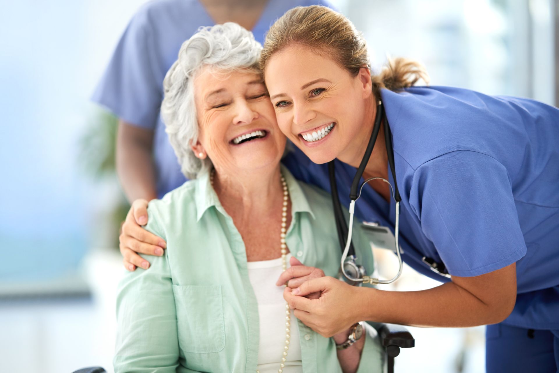 Nurse hugs and smiles with a senior woman in a wheelchair, both smiling. Another nurse is in the background.
