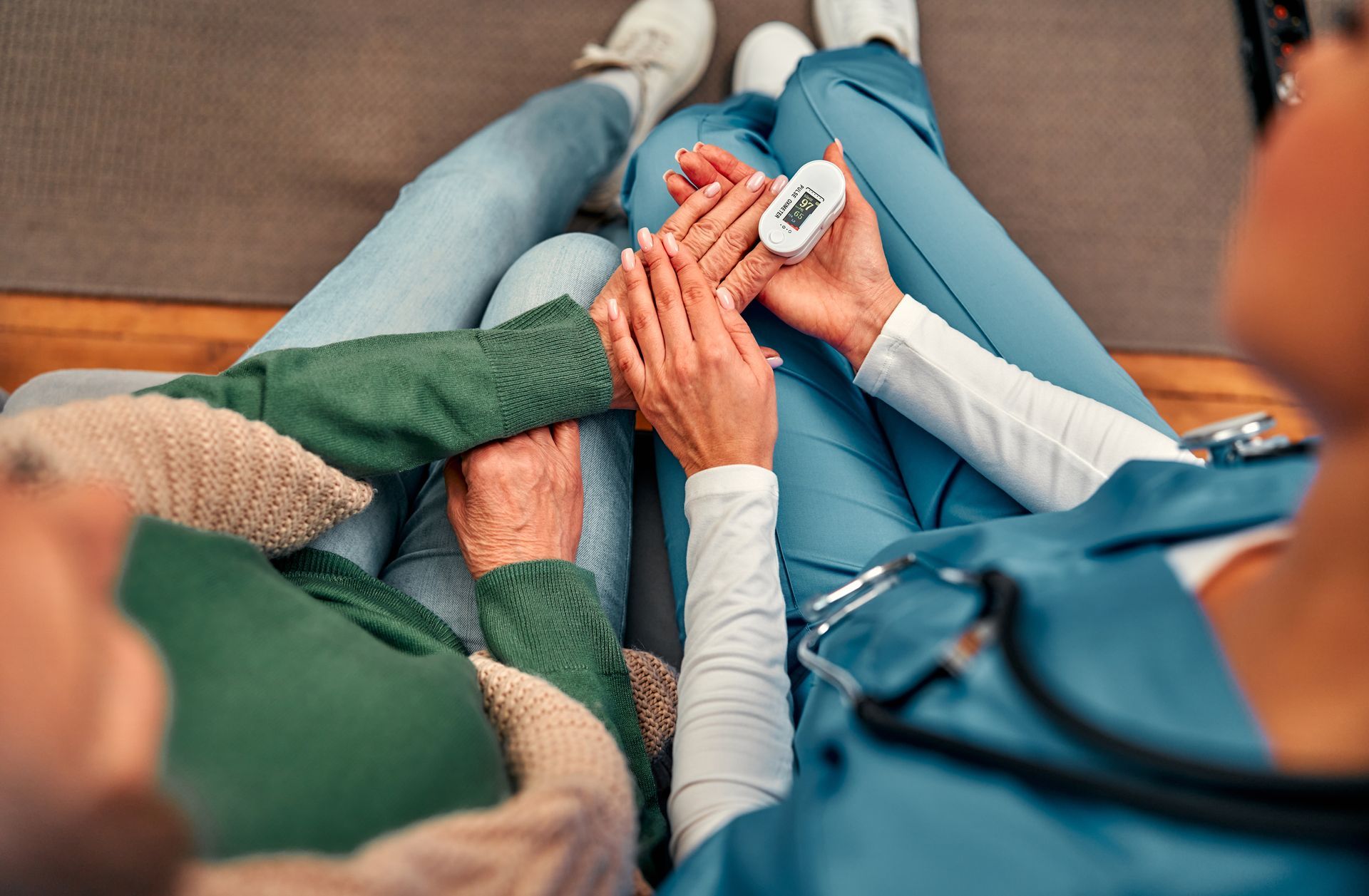 Person's finger in a pulse oximeter by a healthcare worker. Both are seated on a couch.