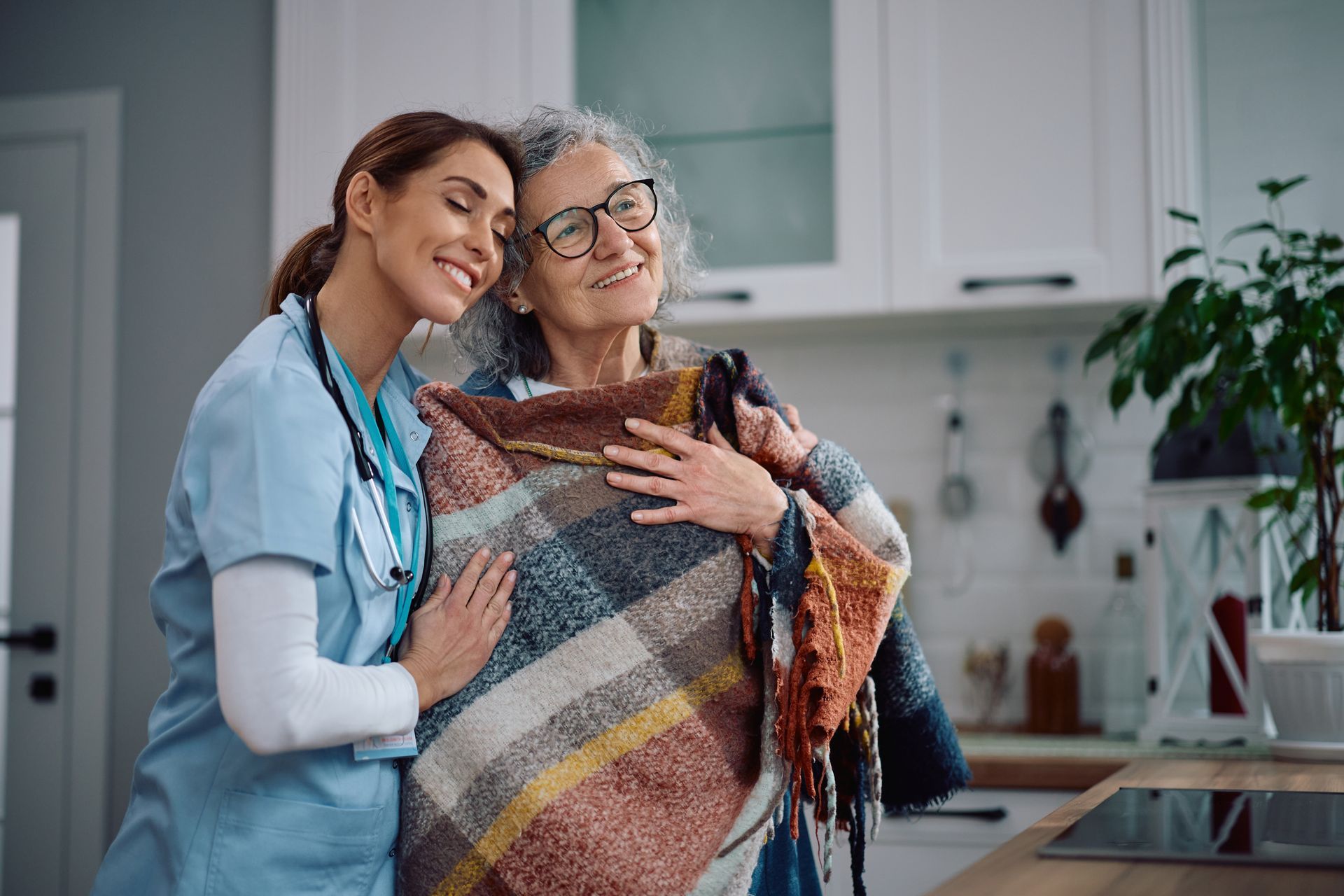 A caregiver hugs an elderly person wrapped in a blanket in a kitchen, both smiling.