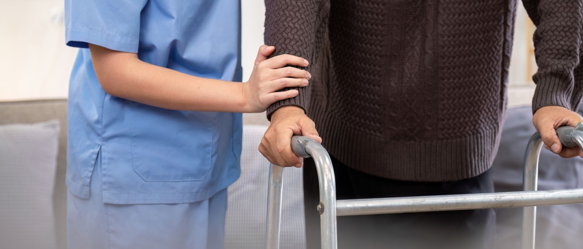 Caregiver assisting a person using a walker, indoors. Blue scrubs, brown sweater.