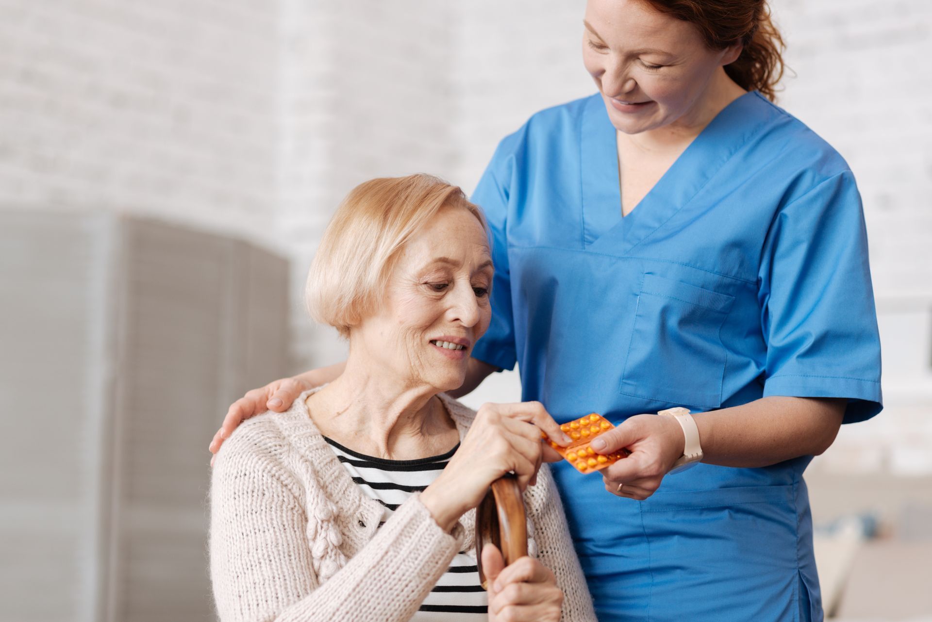 Nurse in blue scrubs gives pills to elderly person holding a cane.