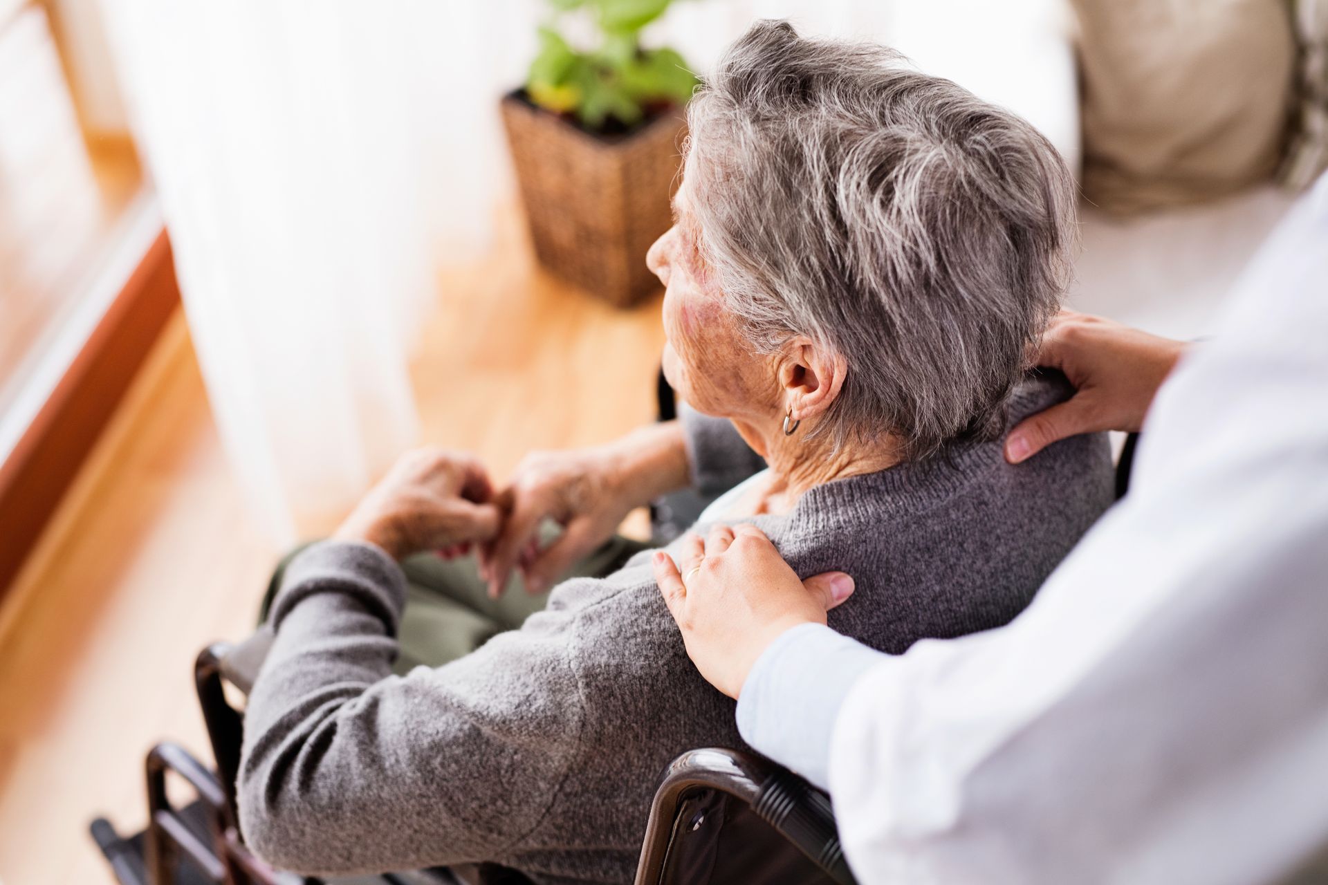 Elderly person in wheelchair with caregiver's hands on shoulders, indoors.