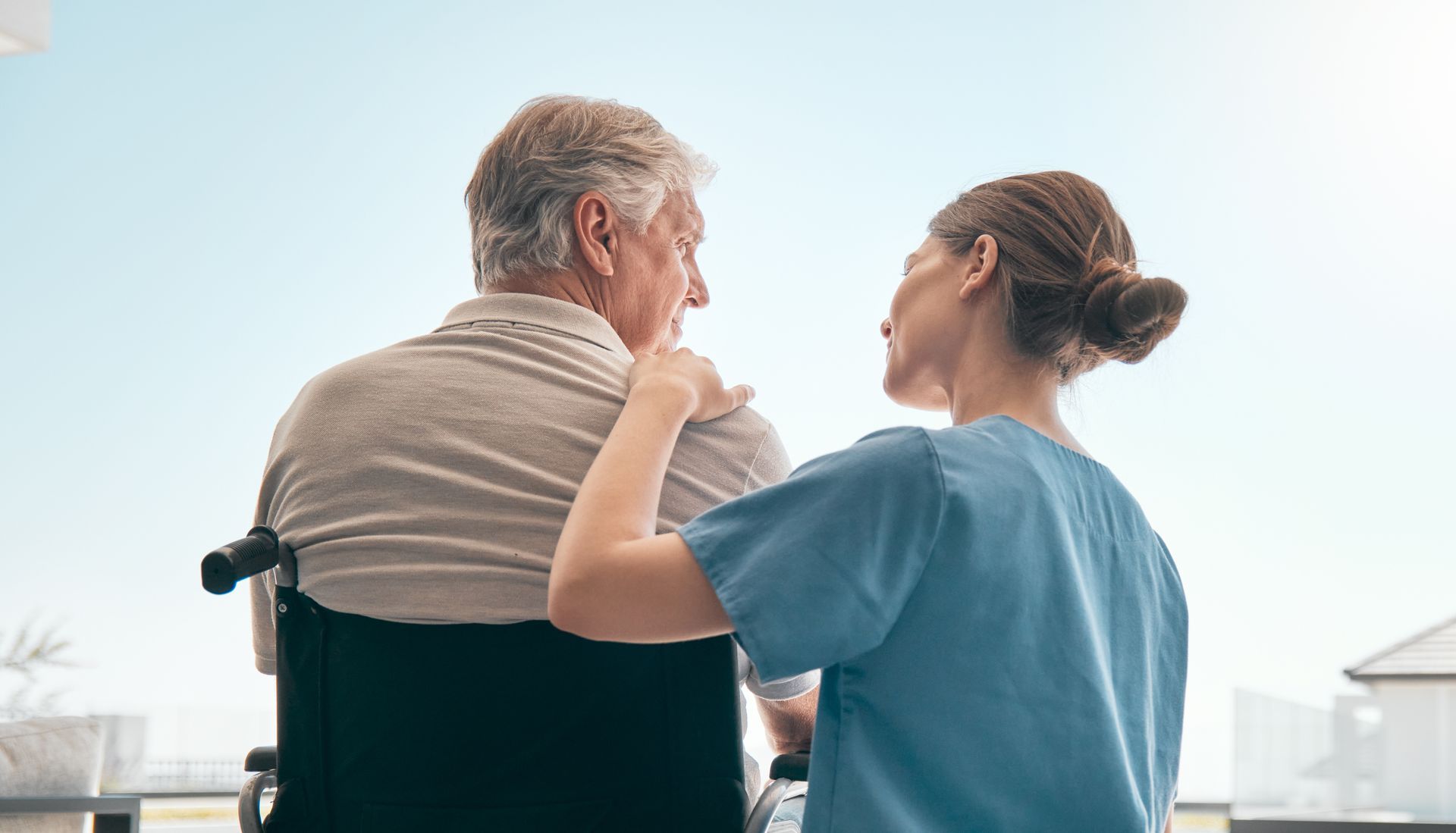 A person in a wheelchair and a caregiver looking at each other outside. The caregiver has their hand on the other person's shoulder.