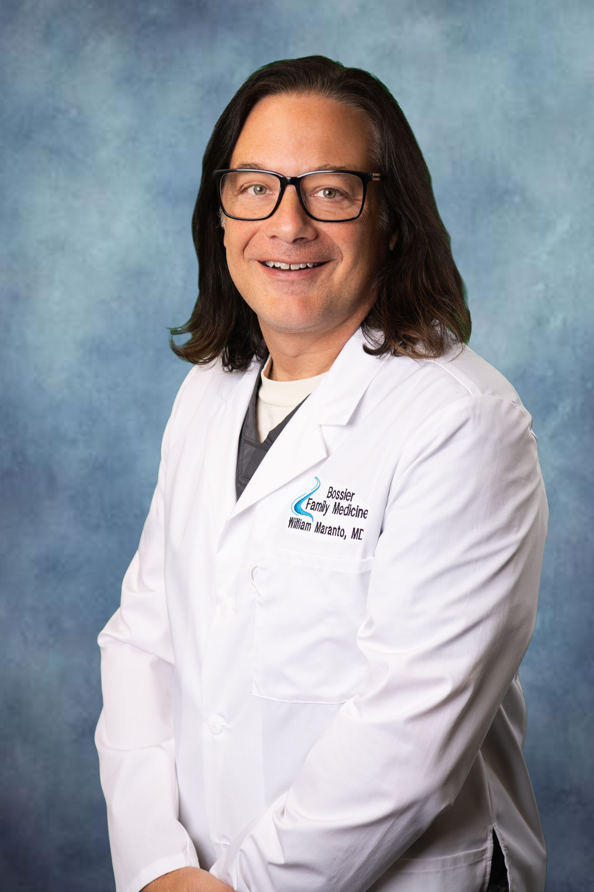 A man in a white lab coat and glasses is standing in front of a blue background.