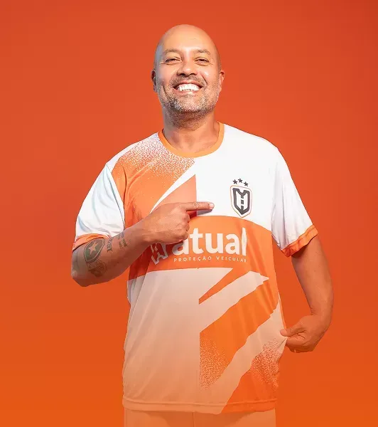 Homem sorridente com camisa laranja e branca apontando para o peito contra um fundo laranja.