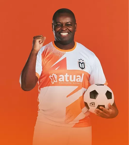 Jogador de futebol com camisa laranja e branca sorrindo, segurando uma bola e erguendo o punho contra um fundo laranja.
