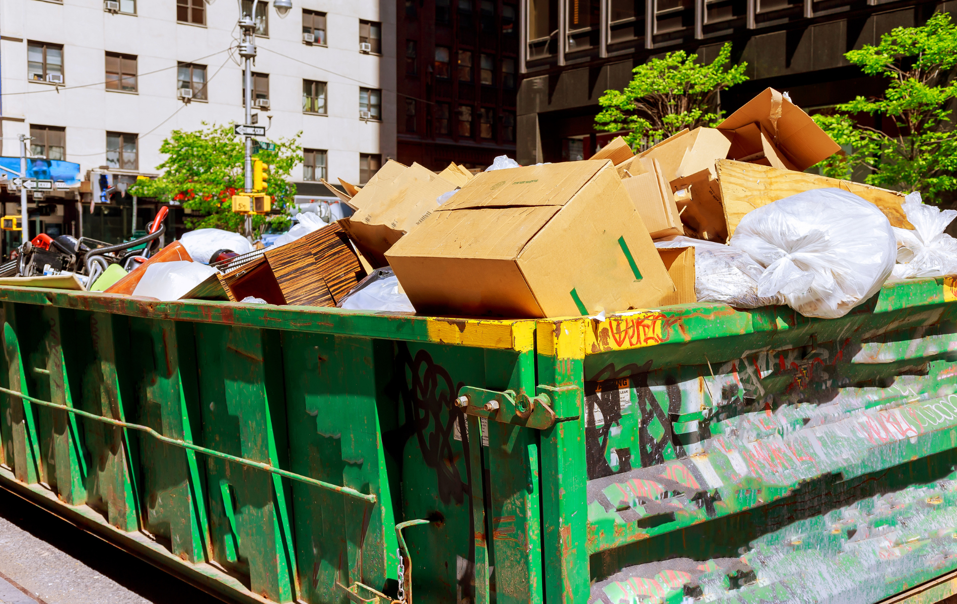 Green dumpster overflowing with cardboard boxes and trash on a city street.