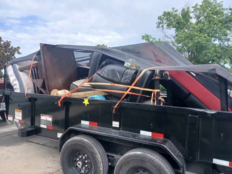 Black trailer loaded with discarded furniture and debris, secured with orange straps.
