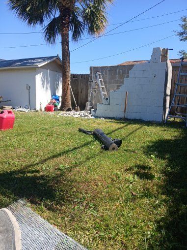 Backyard with construction: partially built concrete wall, ladder, tools, palm tree, and shed.
