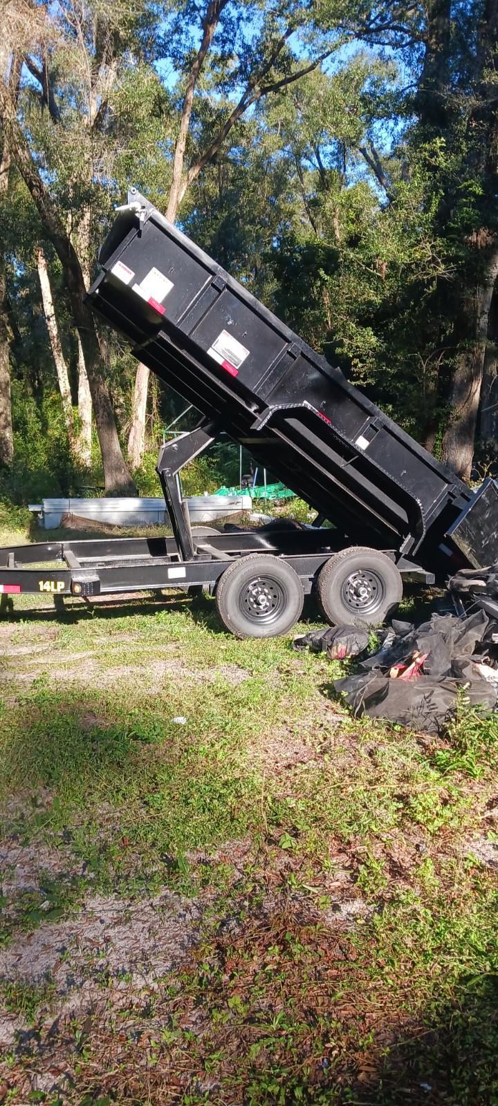 Black dump trailer tilted up, unloading debris onto grass near trees.