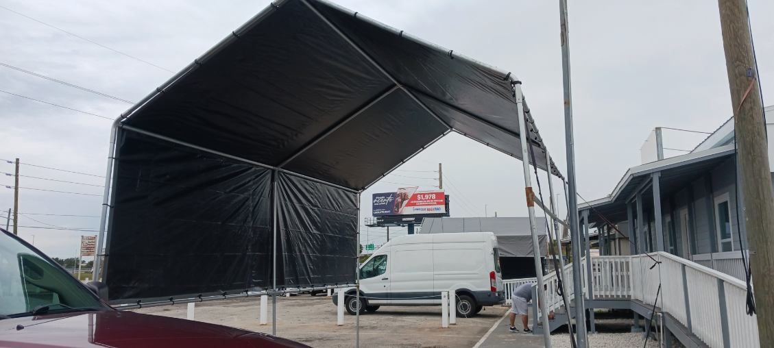 Black tent structure over a white van and a building with a ramp. Overcast day.
