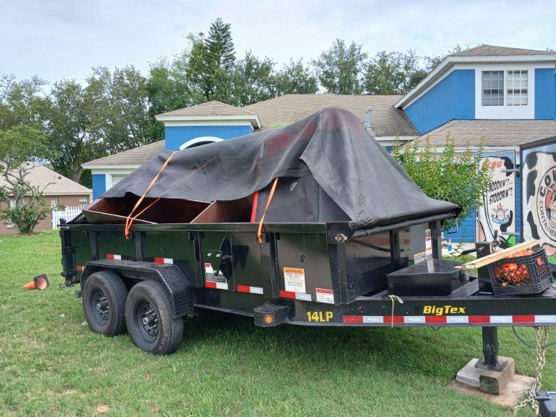 Black trailer with cardboard and tarp secured by straps, on green grass. House in background.