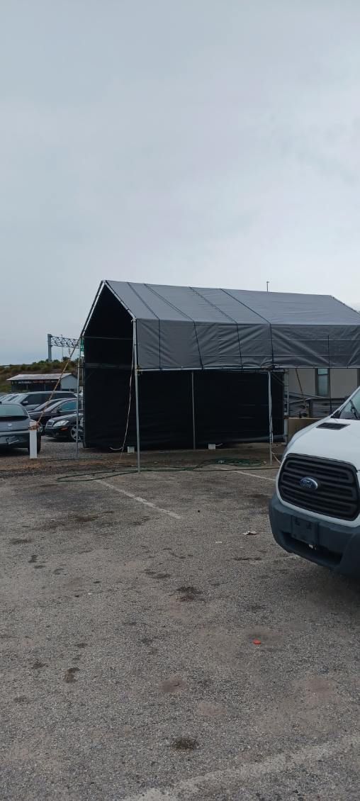 A dark shed with a gray tarp roof and a white van parked in a gravel parking lot.