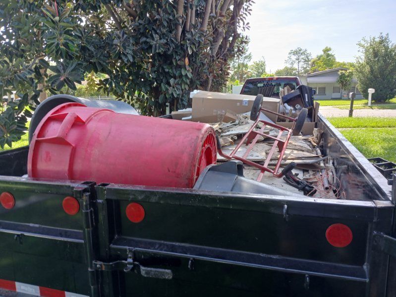 A black truck bed overflowing with trash, including a red barrel and metal scraps, parked on grass.