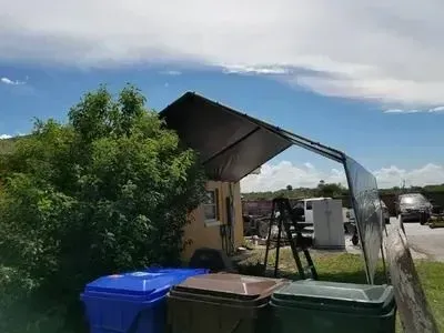 A tarp-covered structure with trash bins in front, against a partly cloudy sky.