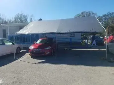 Red car parked under a white tent; other cars and building in the background. Sunny day.