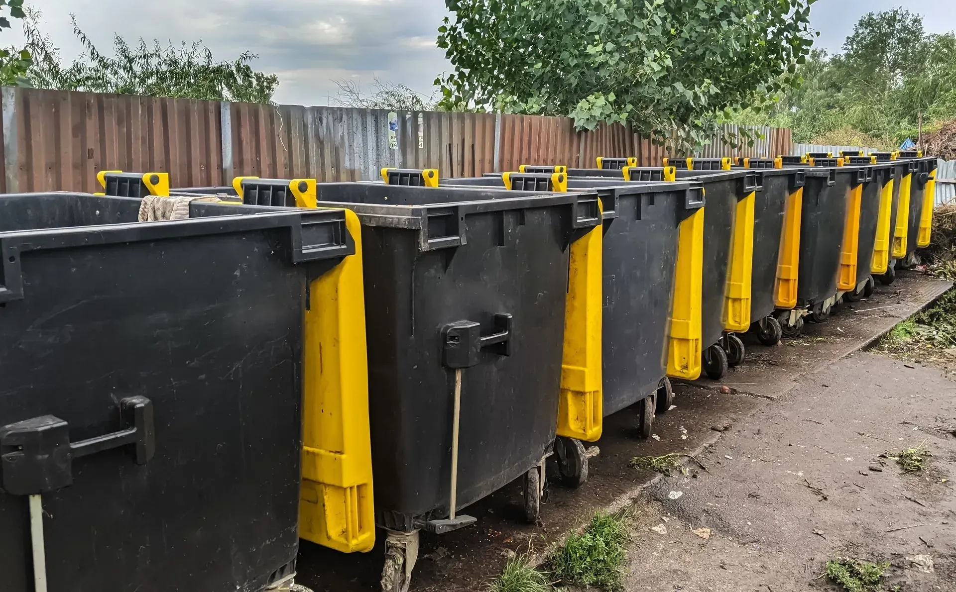 A row of large, black trash dumpsters with yellow accents, lined up along a fence.