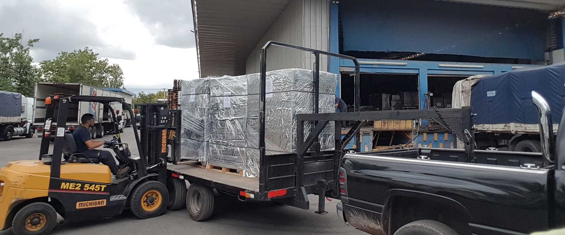 Una carretilla elevadora carga un remolque con cubos de metal en una nave industrial. Hay una camioneta cerca.