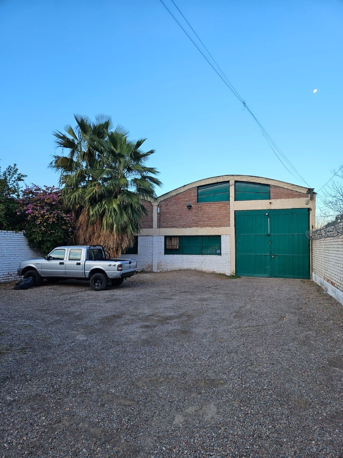 Camioneta plateada estacionada frente a un edificio de ladrillo con puerta verde y palmera bajo un cielo azul.