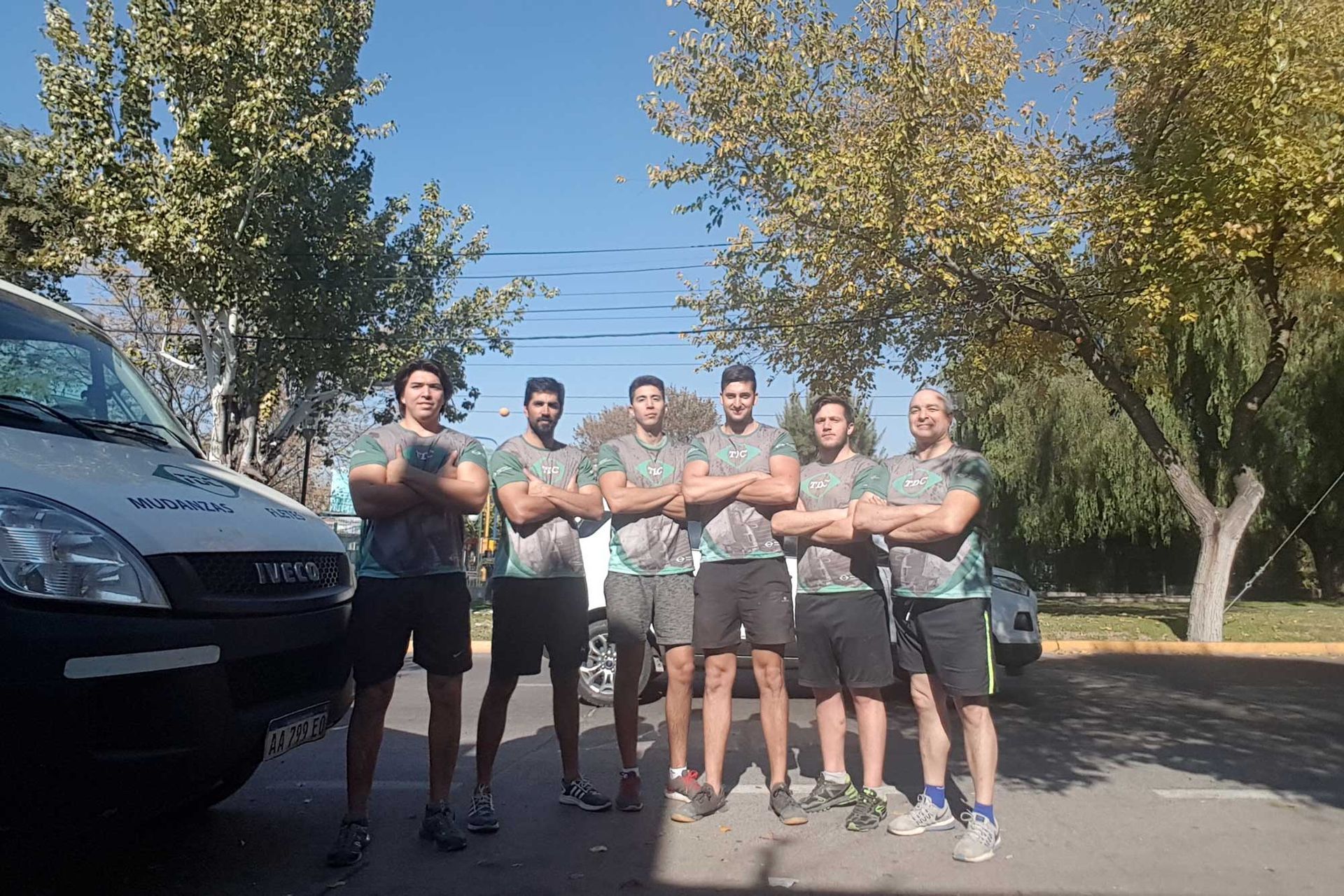 Seis hombres con camisetas y pantalones cortos a juego están de pie con los brazos cruzados cerca de una camioneta al aire libre.