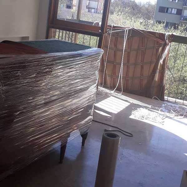 Una cómoda envuelta en un balcón, luz solar, un rollo de film plástico y árboles al fondo.