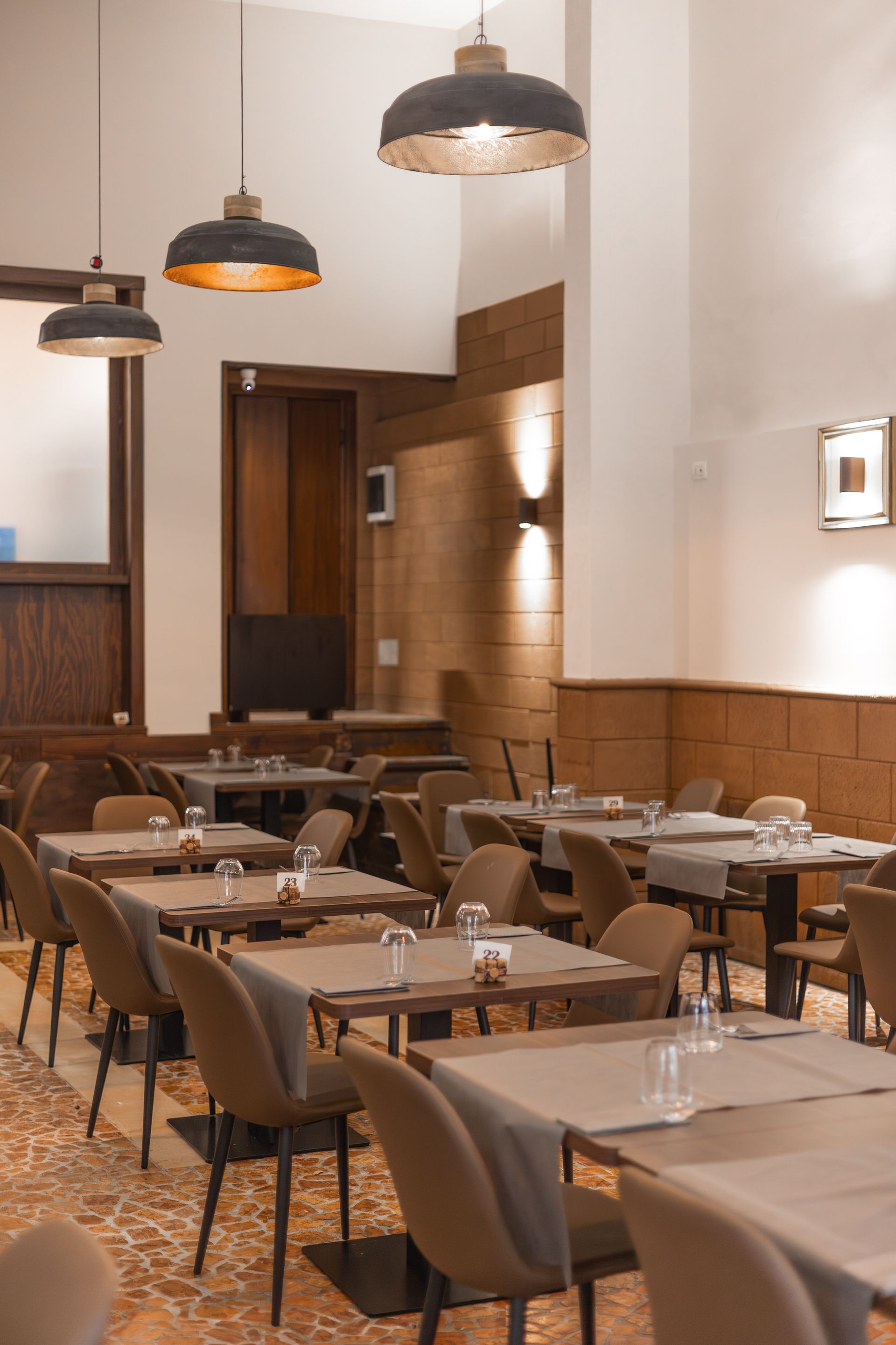 Sala da pranzo di un ristorante moderno con tavoli apparecchiati, sedie grigie, lampade a sospensione e pareti rivestite in legno.