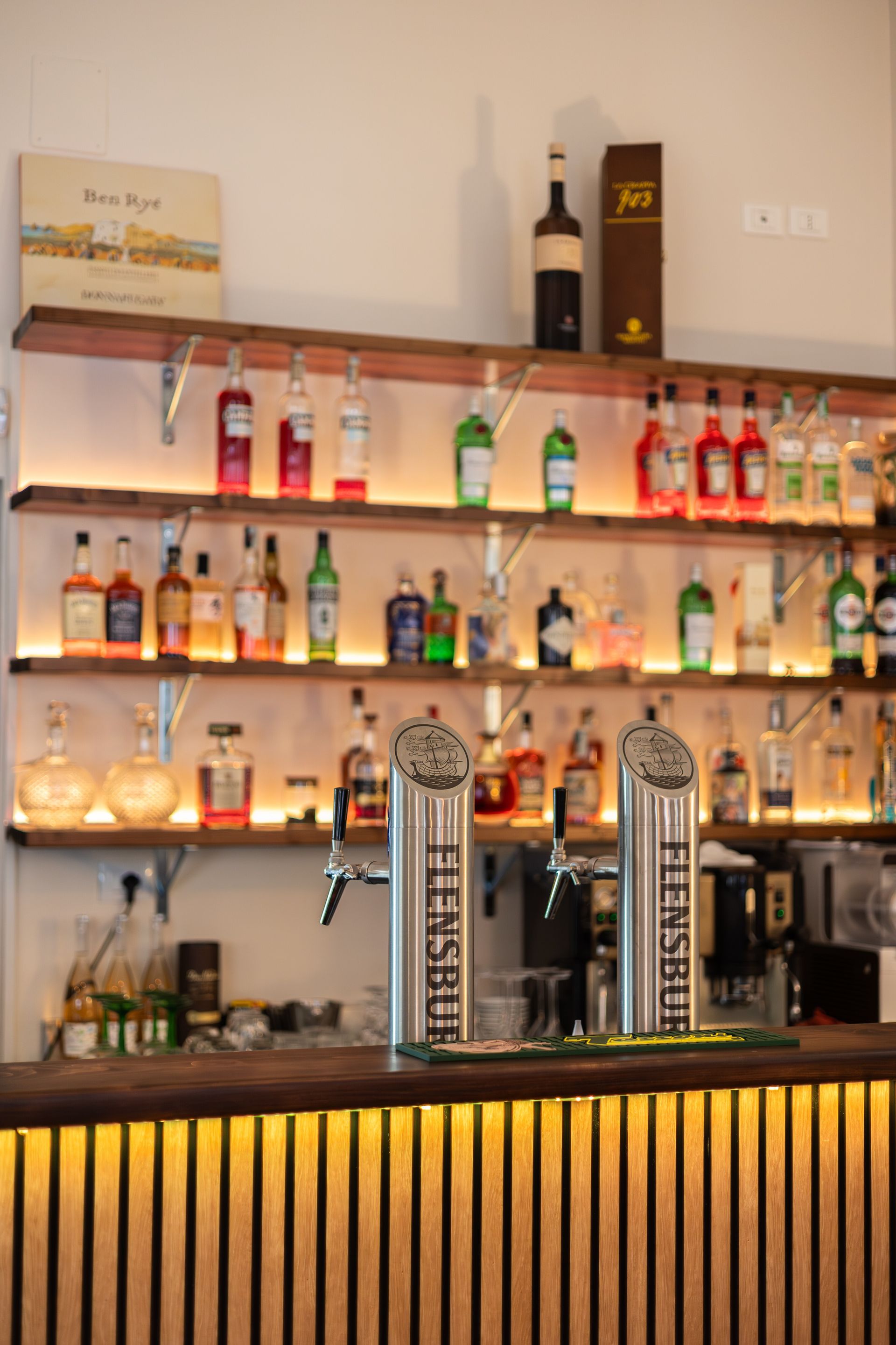 Bancone bar con scaffali illuminati pieni di bottiglie di liquore e due spine di birra dietro un bancone di legno.