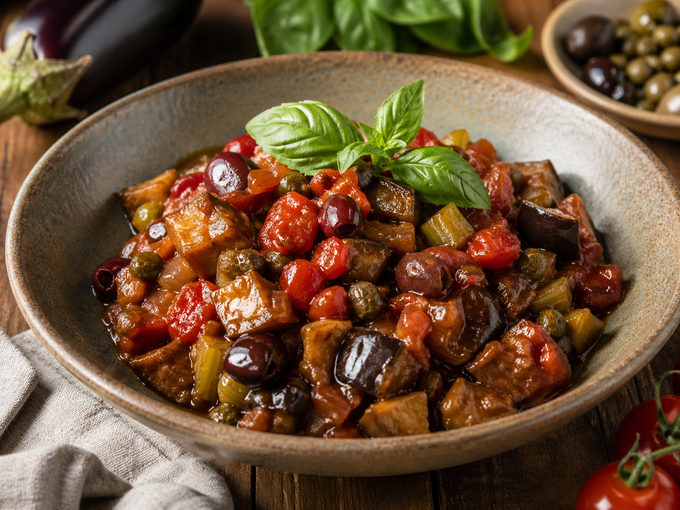 Ciotola di stufato di melanzane con basilico, guarnita con pomodorini e olive su un tavolo rustico.