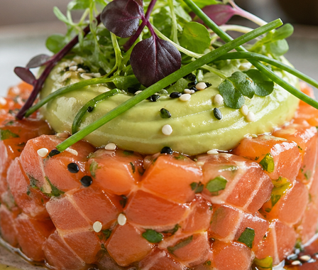 Torretta di tartare di salmone guarnita con crema di avocado, semi di sesamo ed erba cipollina su un piatto bianco.