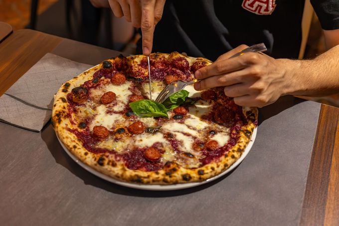 Mani che tagliano una pizza al formaggio e basilico su un tavolo, con due mani che si protendono verso di essa.