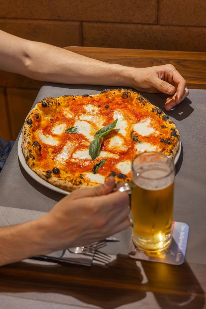 Mani che si allungano per prendere una pizza e una birra su un tavolo in un ambiente simile a un ristorante.