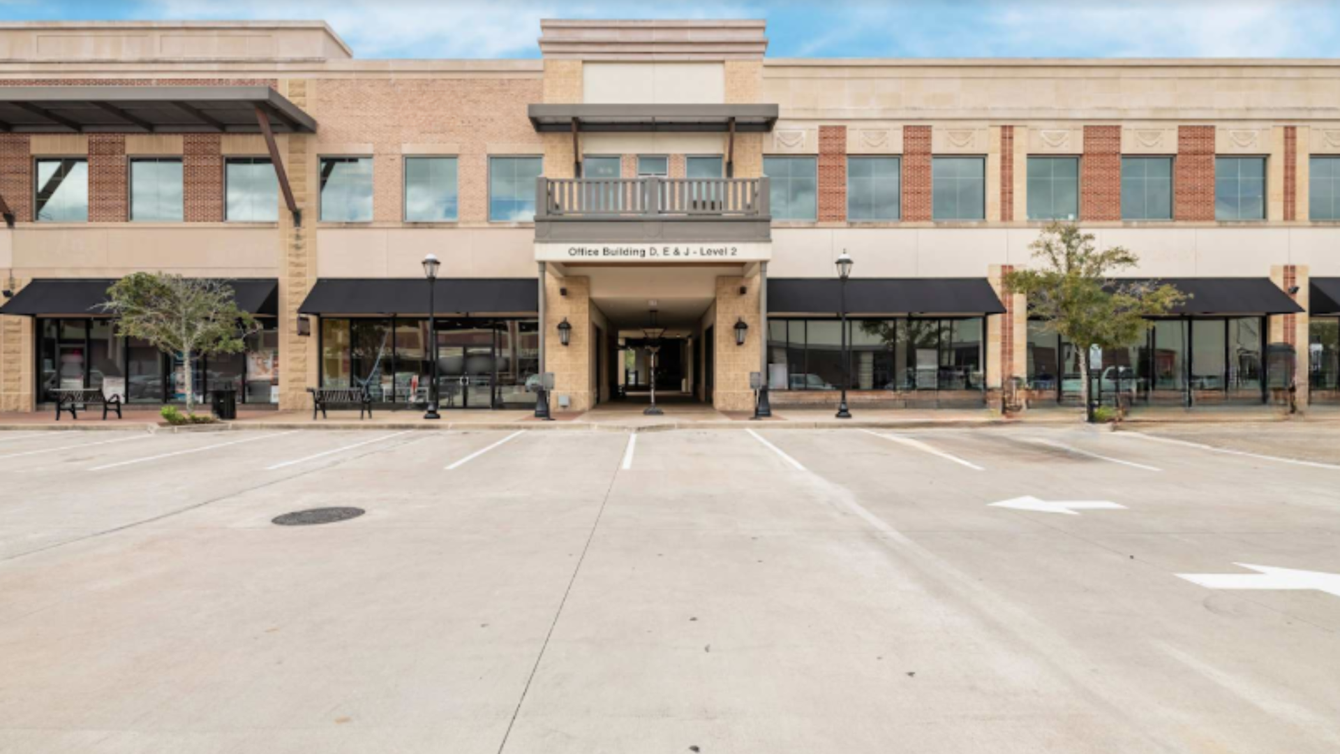 A tan, two-story commercial building with black awnings, a center entryway, and a wide, empty concrete parking lot.