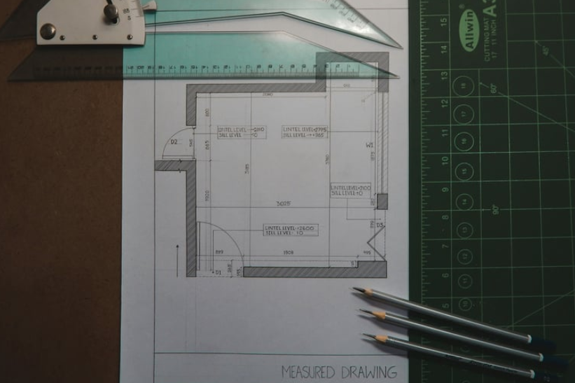 A floor plan sketch on paper with drafting tools, including a ruler and three pencils, on a green cutting mat.