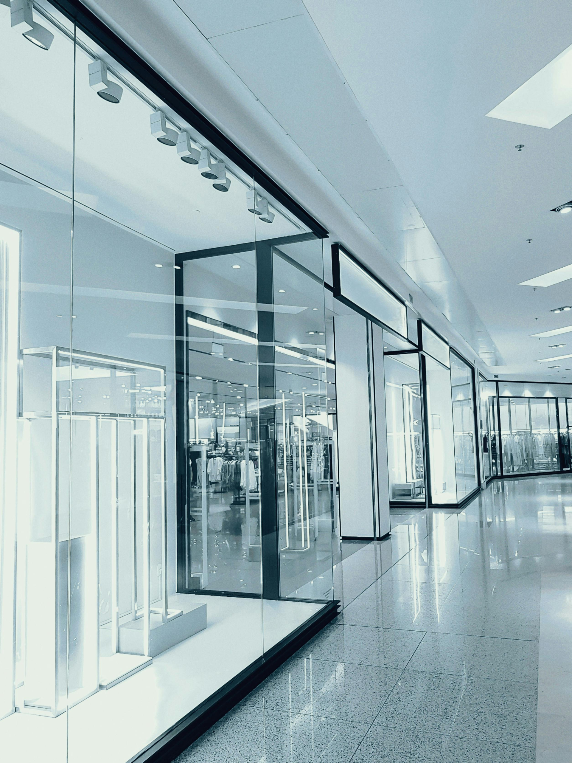 A brightly lit, modern shopping mall corridor with glass storefronts and polished, speckled flooring.