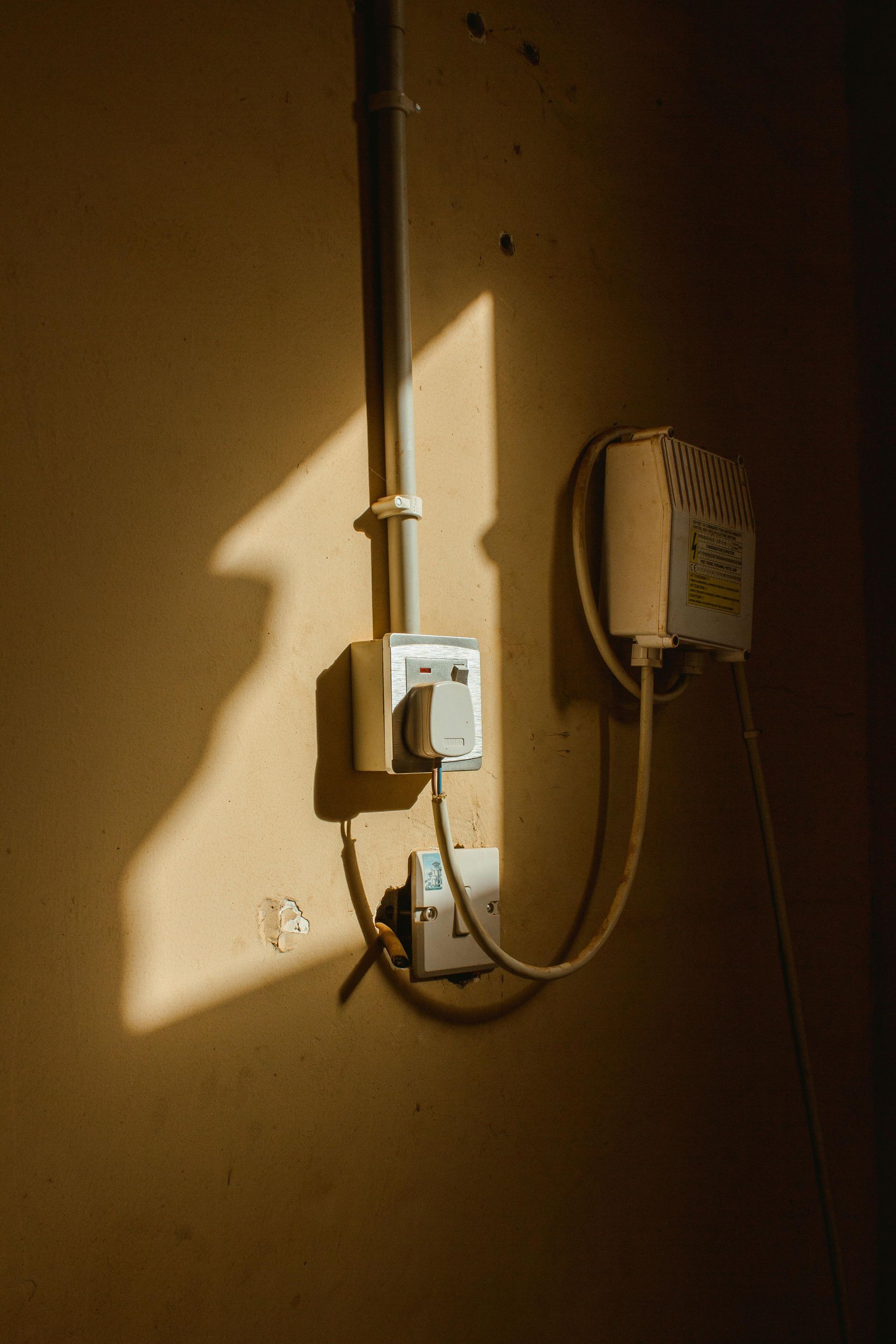A wall with an electrical conduit, two white wall sockets, and a connected device, lit by a strong shadow.