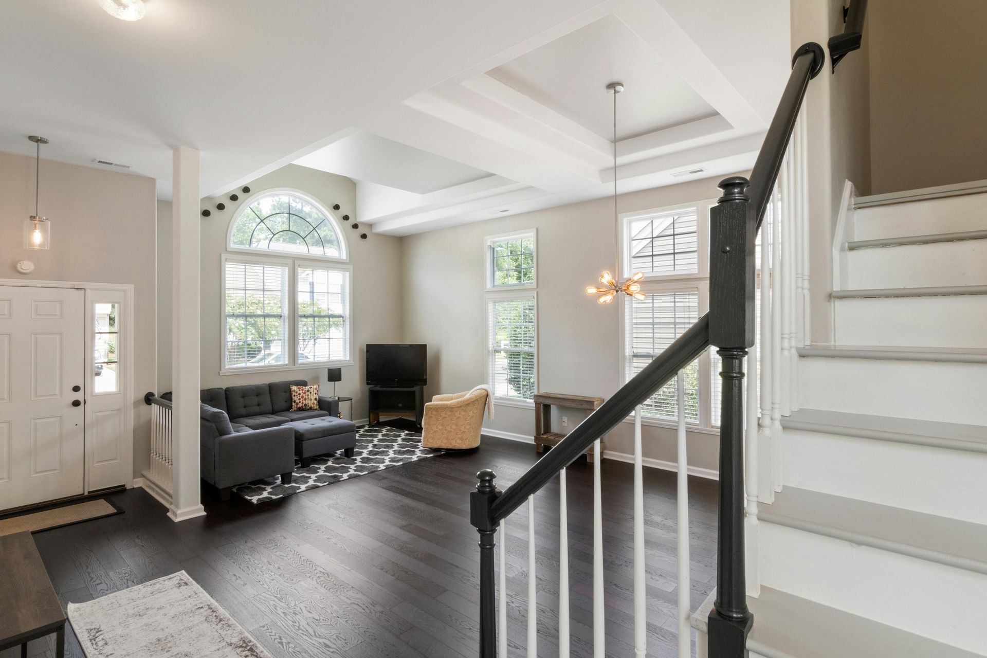 A living room with dark hardwood floors, a gray sectional sofa, a tan armchair, large windows, and a white staircase.